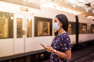 Genç Latin kadın yüz maskesi takıyor ve Coronavirus salgını sırasında metro istasyonunda akıllı telefonu kullanıyor..