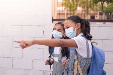 Little girl pointing something to her friend wears a medical mask on the school on the new normal of Coronavirus.