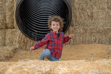 Happy child in red plaid shirt plays in corn pit by hay tunnel