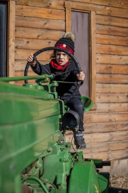 Child in winter hat driving green tractor by wooden barn wall