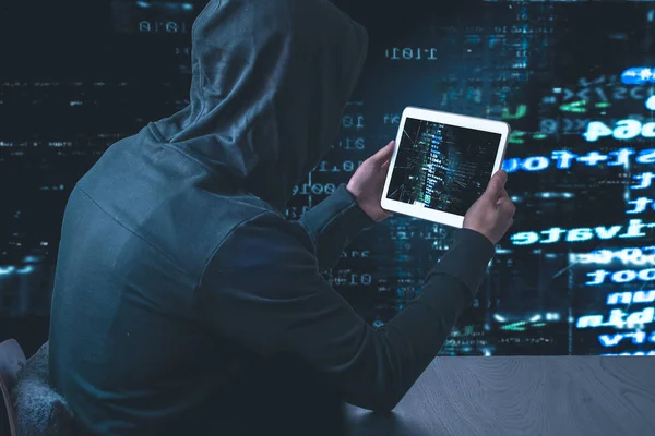 Anonymous hacker in a black hoody with tablet in front of a code background with binary streams cyber security concept