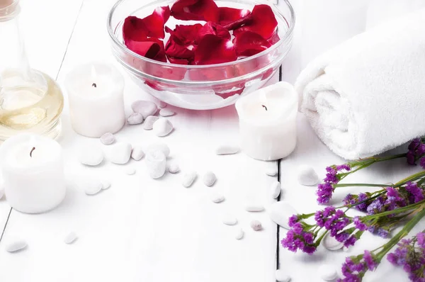 Suyla bir kabın içinde kırmızı bir gül yaprakları, beyaz çakıl ve bir beyaz ahşap arka plan üzerinde beyaz mumlar