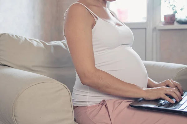 Молодая красивая беременная женщина работает на ноутбуке. Беременная деловая женщина ищет информацию и онлайн-покупки на ноутбуке готовятся к родам и воспитанию на диване дома — стоковое фото