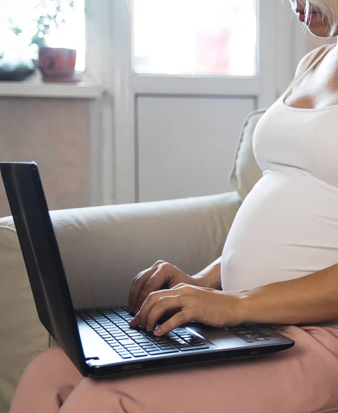 लॅपटॉपवर काम करणारी तरुण सुंदर गर्भवती महिला. लॅपटॉपवर माहिती आणि ऑनलाइन खरेदी शोधत गर्भवती व्यवसाय स्त्री घरी सोफावर जन्म आणि पालकत्व तयार करते — स्टॉक फोटो, इमेज