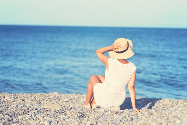 Jonge brunette vrouw in witte zomerjurk zitplaatsen op het strand en op zoek naar de zee. Kaukasische meisje ontspannen en genieten van de rust op vakantie. — Stockfoto