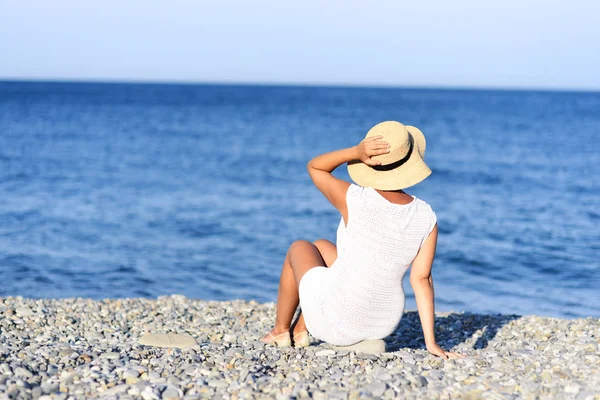 Jonge brunette vrouw in witte zomerjurk zitplaatsen op het strand en op zoek naar de zee. Kaukasische meisje ontspannen en genieten van de rust op vakantie. — Stockfoto