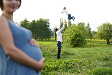 Aile. Aile Portresi. Genç mutlu Aile açık havada yürüyor. Hamile kadın, koca ve çocuk. Baba oğlunu havaya fırlatıyor, ön plandaki anne odak noktasının dışında.