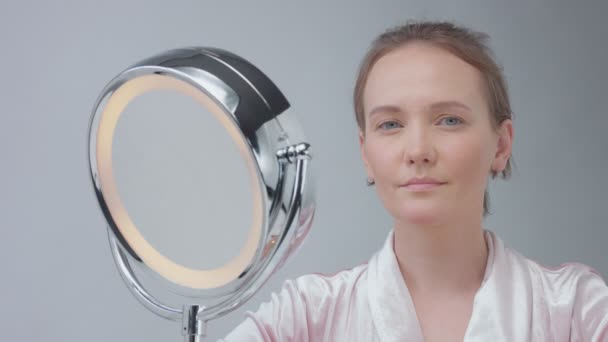 Caucasian woman with mirror with ring light with no makeup look examine her skin and makes facial treatment — Stock Video