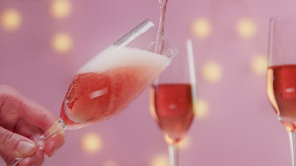 Vino espumoso rosa con mano de mujer y copas sobre fondo rosa con luces navideñas — Vídeos de Stock