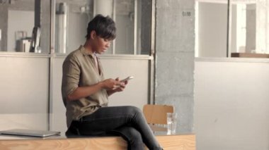 Genç melez ırk siyah kadın masanın üstünde oturuyor ve akıllı telefondan mesaj atıyor.