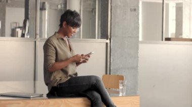 Genç melez ırk siyahi kadın masanın üstünde oturuyor ve akıllı telefondan mesaj atıyor.