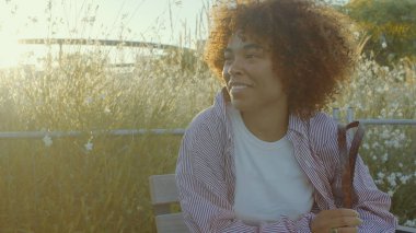 Melez ırk siyah kadın güneş ışığında parkta oturuyor.