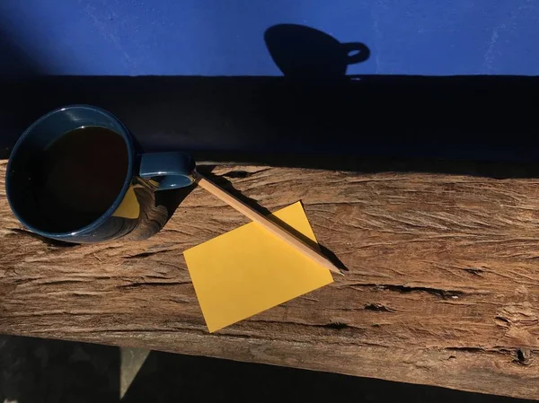 青い壁のホット コーヒー 黄色のメモ帳 その影で自然な茶色の木製のベンチに木製の鉛筆のブルーのカップを反映します — ストック写真