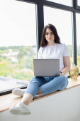 Egy fiatal nő ül az ablak mellett laptoppal. Otthoni munka.