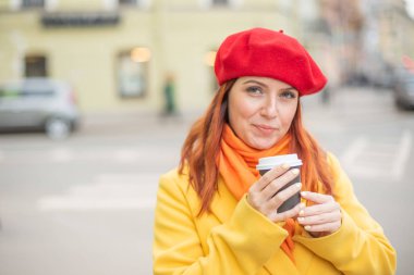 Kızıl saçlı genç kadın bir sarı ceket ve kırmızı bere sıcak tutmak için sokakta kahve içiyor