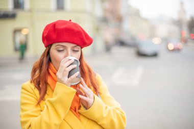Kızıl saçlı genç kadın bir sarı ceket ve kırmızı bere sıcak tutmak için sokakta kahve içiyor