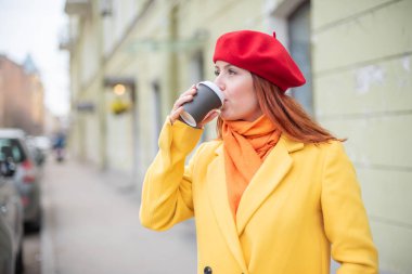 Kızıl saçlı genç kadın bir sarı ceket ve kırmızı bere sıcak tutmak için sokakta kahve içiyor