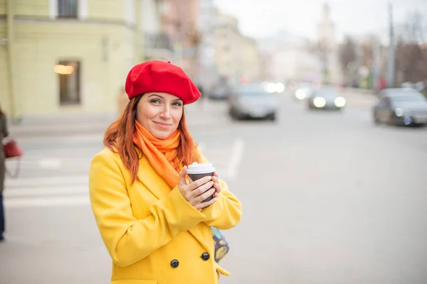 Die rothaarige junge Frau in gelbem Mantel und roter Baskenmütze trinkt auf der Straße Kaffee, um sich warm zu halten — Stockfoto