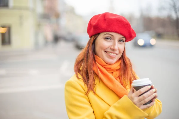 The red-haired young woman in a yellow coat and red beret is drinking coffee on the street to keep warm — Stock Photo, Image