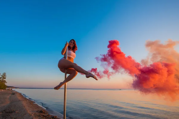 Menina atlética subiu em um pilão portátil na praia e segura uma granada de fumaça vermelha. Areia, pôr do sol, praia . — Fotografia de Stock