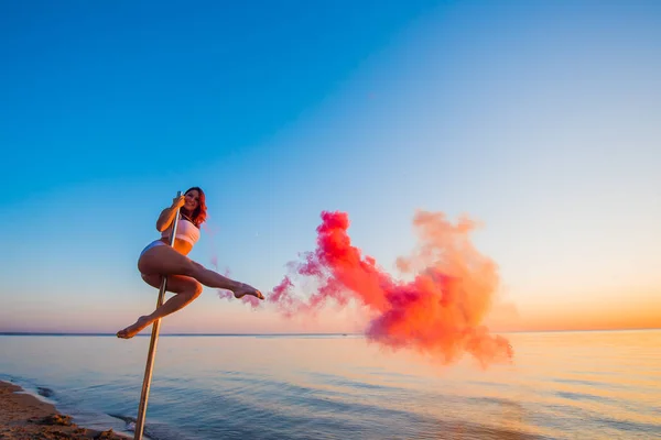 Athlétique fille grimpé sur un pylône portable sur la plage et tient une grenade à fumée rouge. Sable, coucher de soleil, plage . — Photo