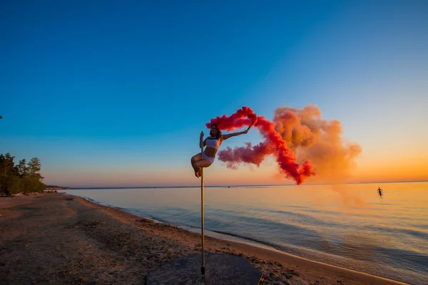 Athlétique fille grimpé sur un pylône portable sur la plage et tient une grenade à fumée rouge. Sable, coucher de soleil, plage . — Photo