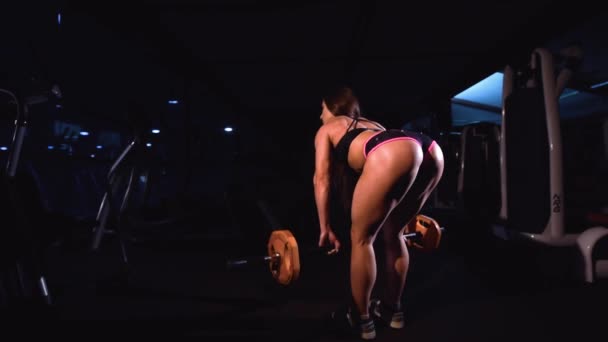 Une fille forte et athlétique en tenue de sport exécute des pistes avec un bar sportif dans une salle de sport sombre. Trains dos et fesses . — Video