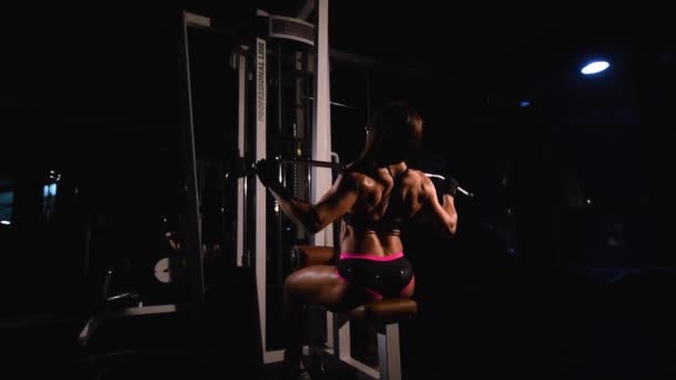 Una chica fuerte, musculosa y atlética, en pantalones cortos, entrena los músculos de la espalda en un simulador horizontal en un gimnasio oscuro . — Vídeos de Stock