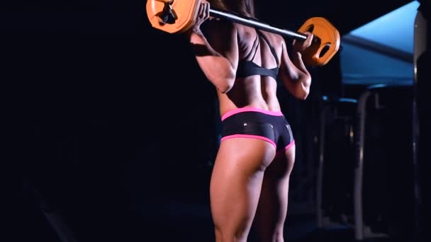 En stark, atletisk flicka i sportkläder utför backarna med en sportbar i ett mörkt gym. Tåg tillbaka och skinkor. — Stockvideo