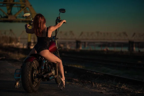 Рыжая женщина в сексуальном белье на высоких каблуках сидит на мотоцикле. Привлекательная рыжеволосая девушка садится на мотоцикл на закате — стоковое фото