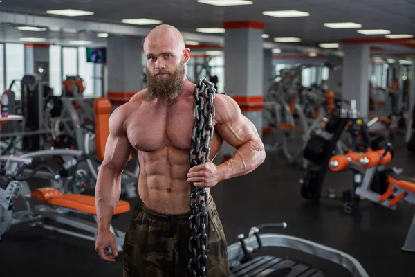 En skallig, skäggiga bodybuilder med stora lättnad muskler har en tung tjock kedja i händerna. Ljust gym. — Stockfoto