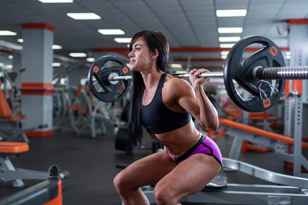 Ein kräftiges, muskulöses dunkelhaariges Mädchen macht Kniebeugen mit einer hellen Langhantel. Fitnessstudio — Stockfoto