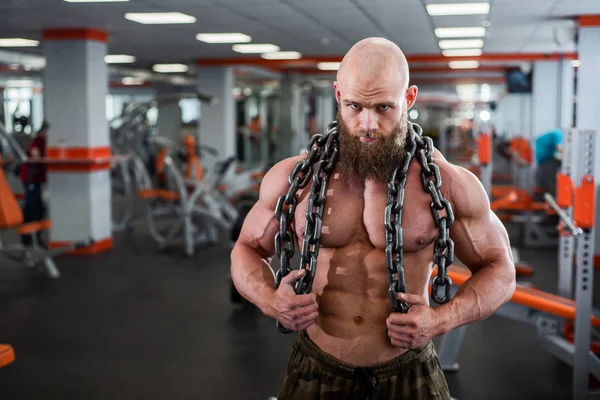 Een kale, bebaarde bodybuilder met grote reliëf spieren heeft een zware dikke ketting in zijn handen. Lichte fitnessruimte. — Stockfoto