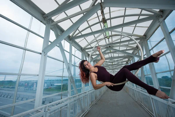 La gymnaste aérienne aux cheveux roux dans un combo sportif se produit au cerceau aérien dans un passage pour piétons . — Photo