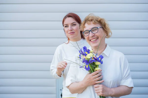 Adult daughter gives flowers to her mother outside, in the courtyard of the house. Spending time together, celebrating at home on weekends. Mothers Day. Warm intergenerational relationships