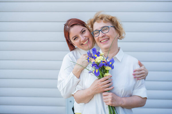 Взрослая дочь дарит цветы своей матери на улице, во дворе дома. Проводим время вместе, празднуем дома по выходным. День Матери. Теплые отношения между поколениями
