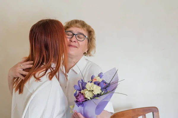 Daughter giving flowers to adult mother. Daughter gives flowers to her mother and hugs her. Spending time together, celebrating at home on weekends. Mothers Day