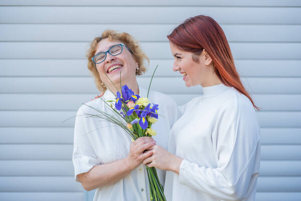 Взрослая дочь дарит цветы своей матери на улице, во дворе дома. Проводим время вместе, празднуем дома по выходным. День Матери. Теплые отношения между поколениями
