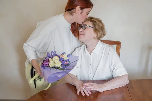 Daughter giving flowers to adult mother. Daughter giving flowers to mother and kissing her on the cheek. Spending time together, celebrating at home on weekend