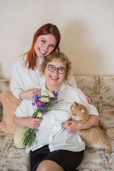 Filha dá flores para uma mãe adulta sentada no sofá na sala de estar. Passar tempo juntos, celebrar em casa aos fins-de-semana. Dia das Mães. Relações intergeracionais quentes — Fotografia de Stock