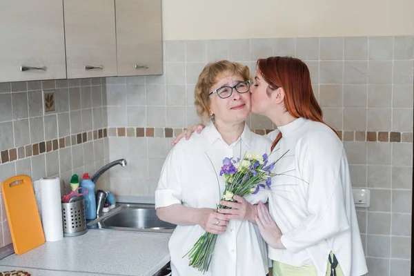 Daughter gives flowers to her adult mother and kisses her in the kitchen. Spending time together, celebrating at home on weekends. Mothers Day. Tears of happiness.