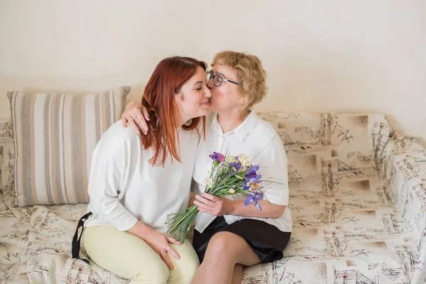 Daughter gives flowers to an adult mother sitting on the sofa in the living room. Spending time together, celebrating at home on weekends. Mothers Day. Warm intergenerational relationships