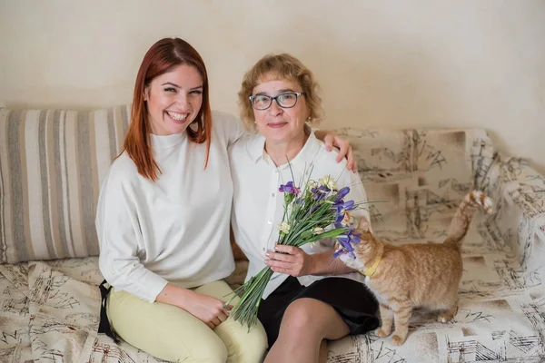 Η κόρη δίνει ένα μπουκέτο σε μια ενήλικη μητέρα που κάθεται σε έναν καναπέ στο σαλόνι. Περνάμε χρόνο μαζί, γιορτάζοντας στο σπίτι τα Σαββατοκύριακα. Ημέρα της μητέρας. Η γάτα τρώει λουλούδια — Φωτογραφία Αρχείου