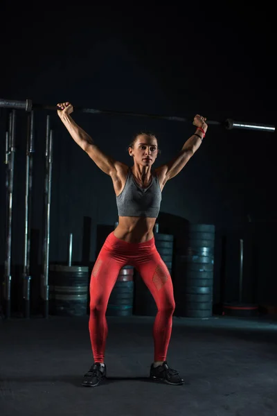 Молодая, европейская, мускулистая девушка в красных леггинсах, делает упражнения с штангой в тренажерном зале для кроссфита. Темный фон — стоковое фото