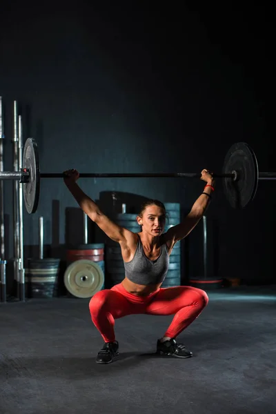 Молодая, европейская, мускулистая девушка в красных леггинсах, делает упражнения с штангой в тренажерном зале для кроссфита. Темный фон — стоковое фото