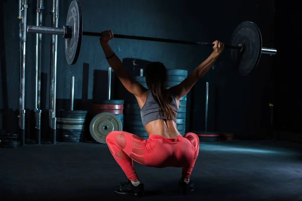 Νέος, Ευρωπαίος, μυώδης κορίτσι με κόκκινα κολάν, κάνοντας άσκηση με ένα μπάρα στο γυμναστήριο για διασταύρωση. Σκούρο φόντο — Φωτογραφία Αρχείου