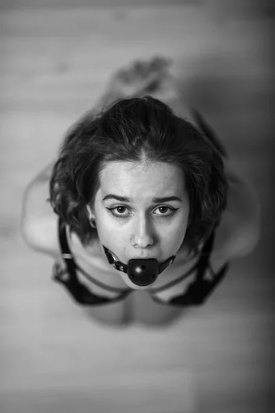 Slavery in society. The girl is on her knees with a gag in her mouth. Sexual bdsm toy. Woman slavery. Slave attire for playing bdsm games. Frightened slave. Black leather bondage with gag. Scared host — Stock Photo, Image