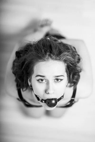 Slavery in society. The girl is on her knees with a gag in her mouth. Sexual bdsm toy. Woman slavery. Slave attire for playing bdsm games. Frightened slave. Black leather bondage with gag. Scared host — Stock Photo, Image