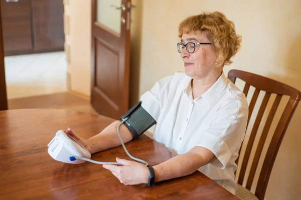 안경을 쓴 노인 여성은 밝은 방에서 전기 장치를 사용하여 혈압을 측정합니다. 흰색 블라우스에 은퇴 한 여자는 테이블에 앉아 집에서 토노미터를 사용 — 스톡 사진