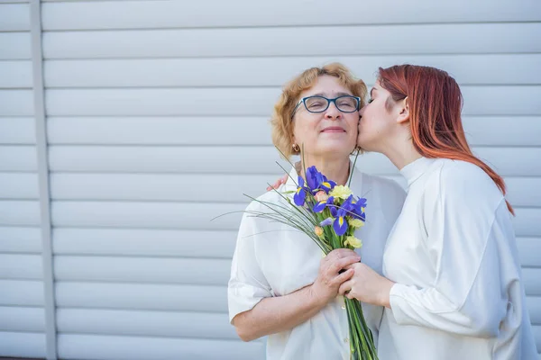 Adult daughter gives flowers to her mother outside, in the courtyard of the house. Spending time together, celebrating at home on weekends. Mothers Day. Warm intergenerational relationships